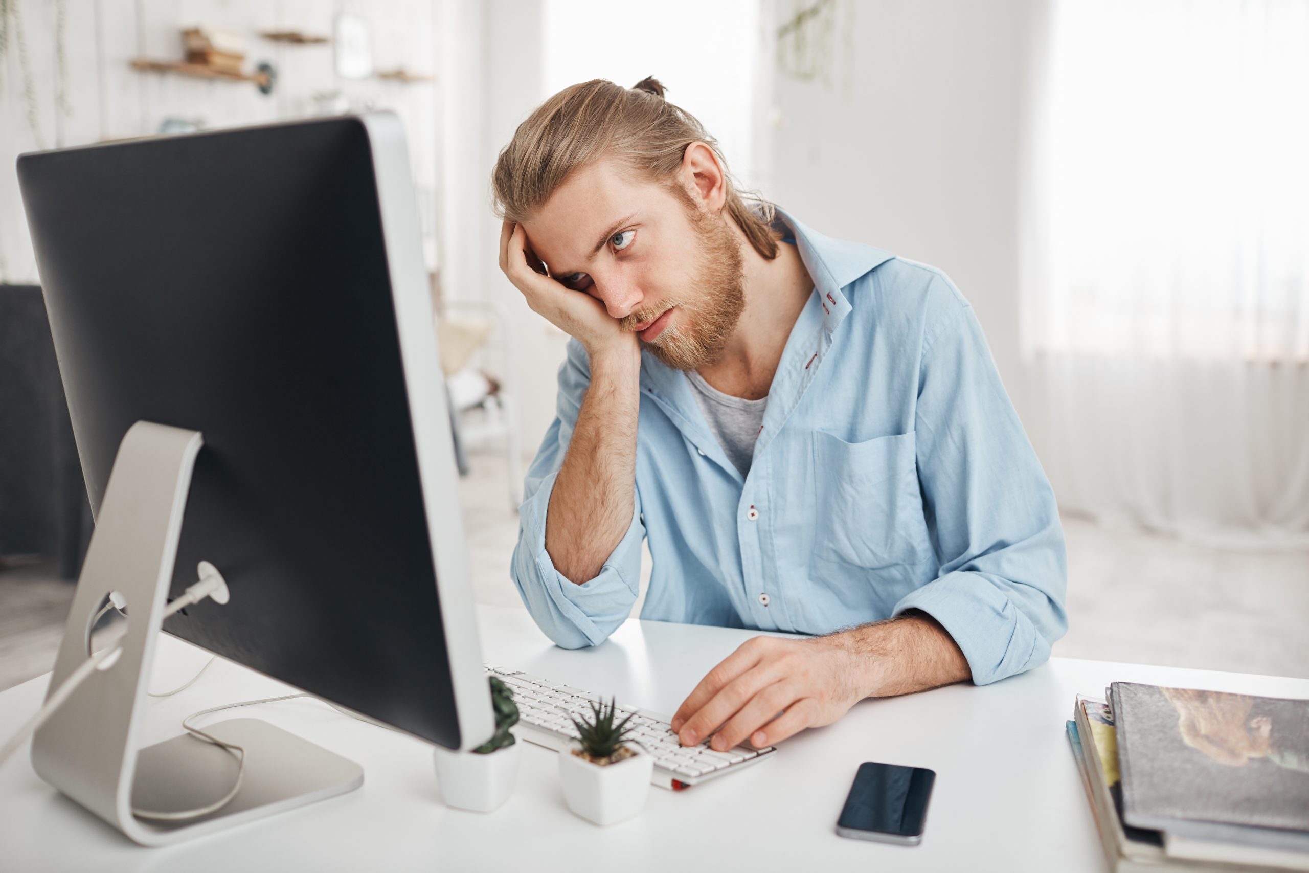 Photo d'un homme derrière son ordinateur tenant sa tête sur sa main d'un air blasé et fatigué par ce qu'il voit sur son écran.